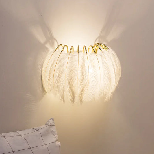 Gray feather wall sconce with a gold metal frame, casting a warm glow on a white wall.
