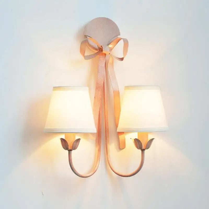 Pink wall sconce with two fabric shades and a prominent ribbon bow detail, mounted on a wall.