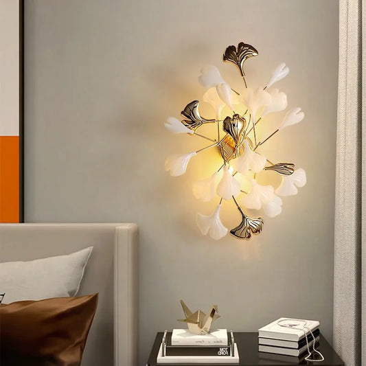 A modern gold wall sconce designed with multiple white ginkgo leaves, casting a warm glow on a neutral wall.