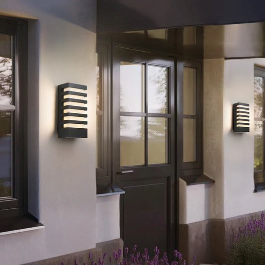 A black modern outdoor wall sconce with horizontal light slats, mounted on a light-colored wall next to a window and dark door.