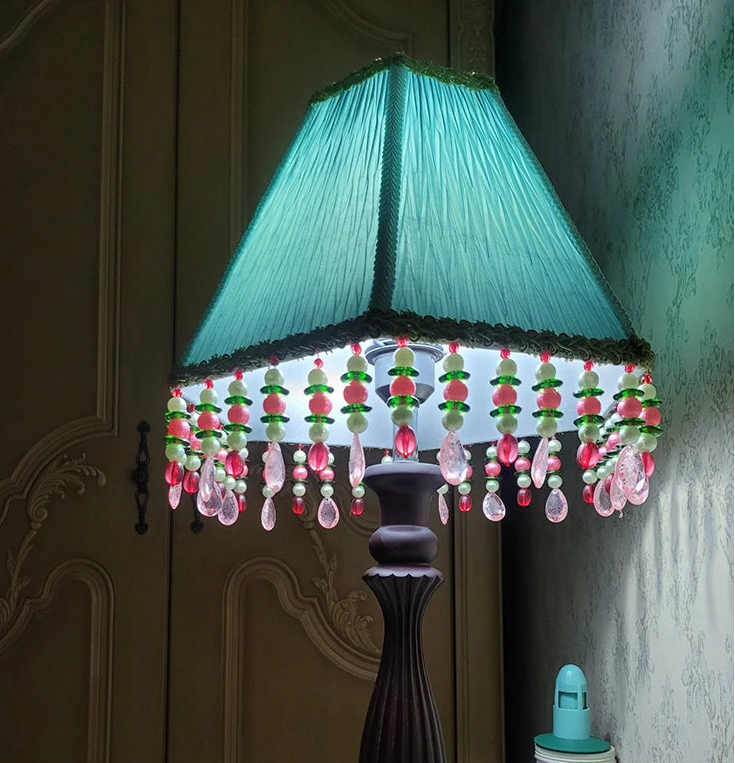 A teal blue pleated fabric empire lampshade with beaded tassels, fitted onto a lamp base in a dimly lit room.
