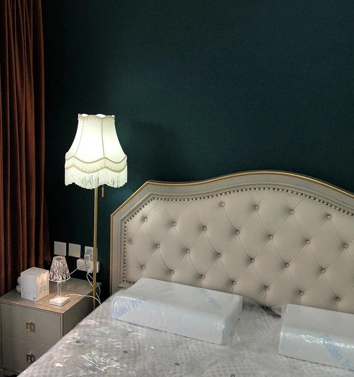 Floor lamp with a cream fabric bell lampshade with fringe next to a bed