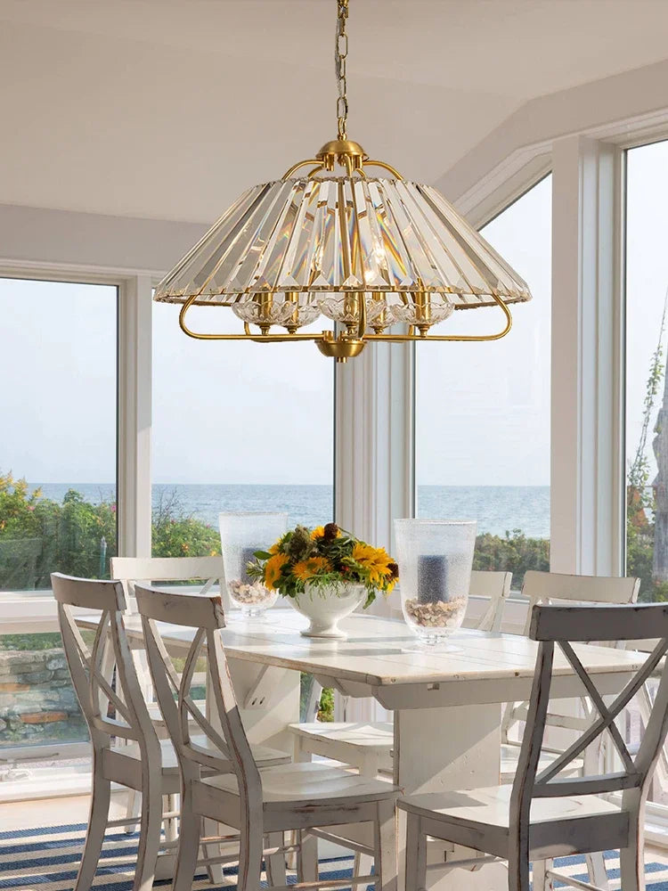 A gold crystal faceted pendant light suspended above a white dining table by a large window overlooking the ocean.