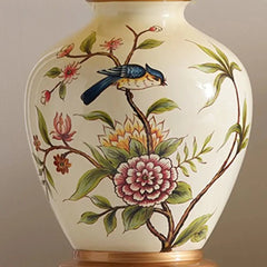 Close-up of the ceramic base of a table lamp, showcasing a detailed painting of a blue bird perched on a branch with pink and yellow flowers.