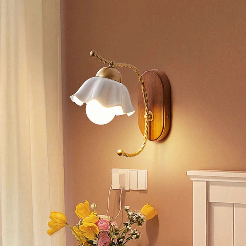 A wall sconce with a white ceramic flower shade and gold arm mounted on a wooden base, illuminated in a room with a brown wall and yellow flowers in the foreground.