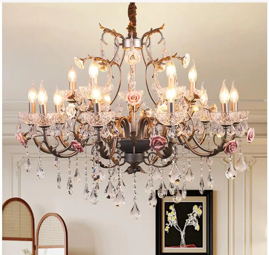 Front view of a decorative gold chandelier featuring intricate metalwork, pink ceramic roses, and clear crystal droplets, illuminating a room.