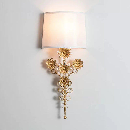 A gold floral crystal wall sconce with a white fabric shade against a white wall.