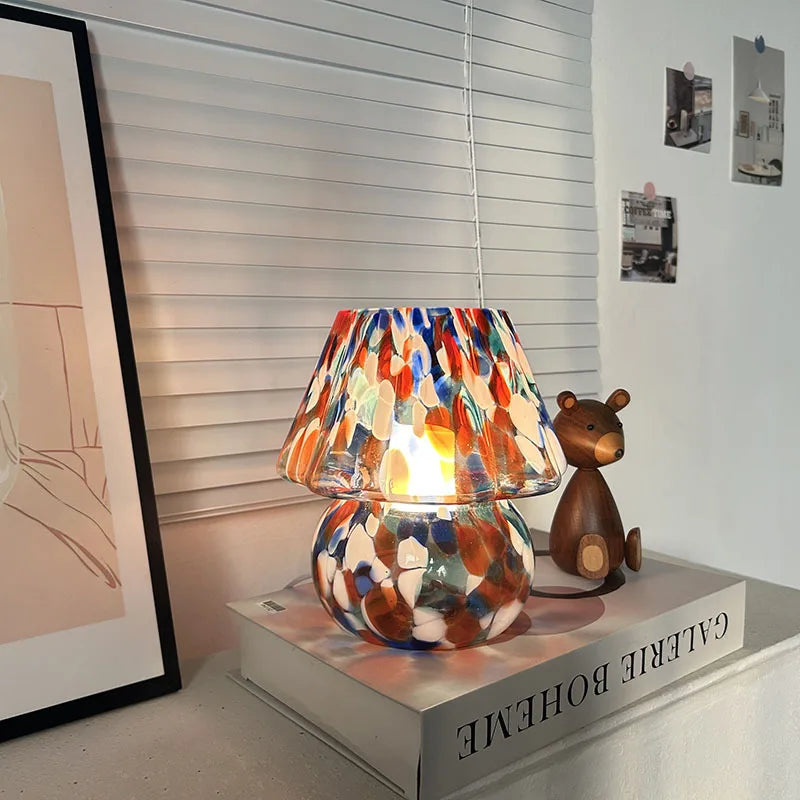 A mushroom-shaped table lamp with a vibrant, multi-colored stained glass shade, illuminated and placed on a book beside a wooden bear.