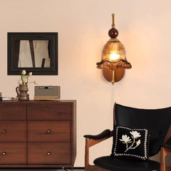 A vintage-style wall sconce with a floral-shaped amber glass shade and a gold arm mounted on a wooden oval backplate, shown in a bedroom setting.