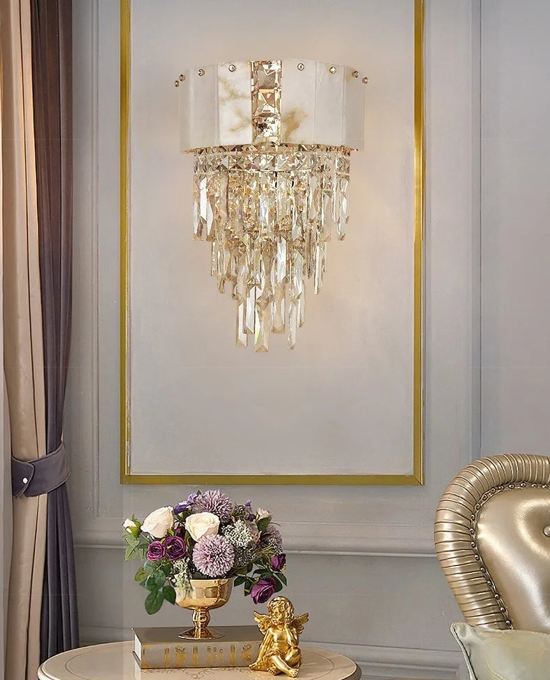 Crystal wall sconce above a table with flowers and a gold statue against a gray wall.