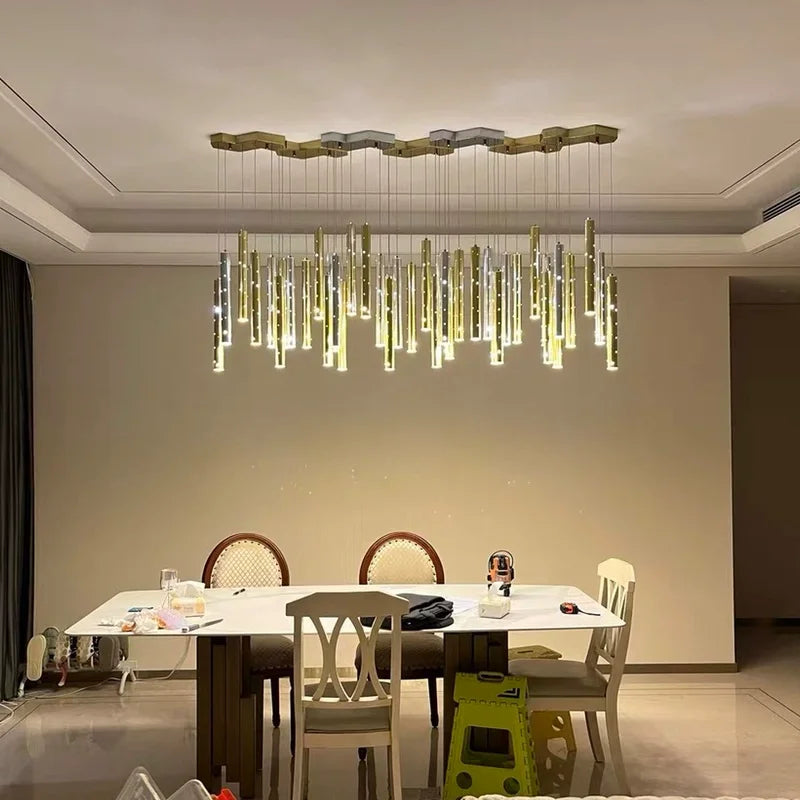 A modern dining room with a large, linear chandelier featuring multiple hanging gold and silver tubes with small light accents, suspended from a hexagonal ceiling plate.