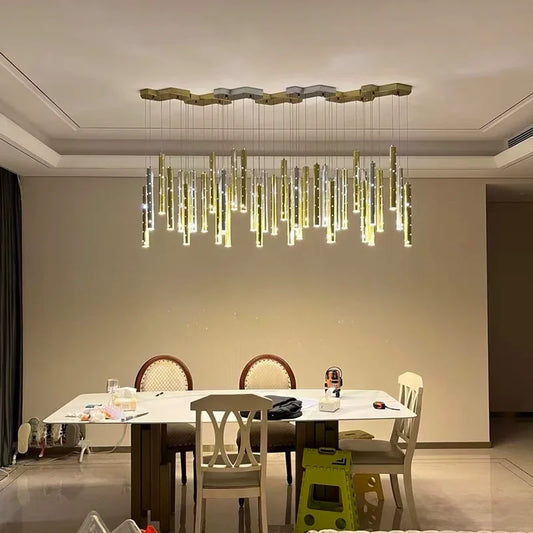 A modern dining room with a large, linear chandelier featuring multiple hanging gold and silver tubes with small light accents, suspended from a hexagonal ceiling plate.