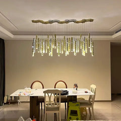A modern dining room with a large, linear chandelier featuring multiple hanging gold and silver tubes with small light accents, suspended from a hexagonal ceiling plate.