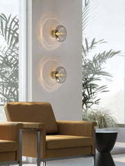 Two modern wall sconces with smoky gray glass diffusers installed on a white wall in a room with large windows and natural light.