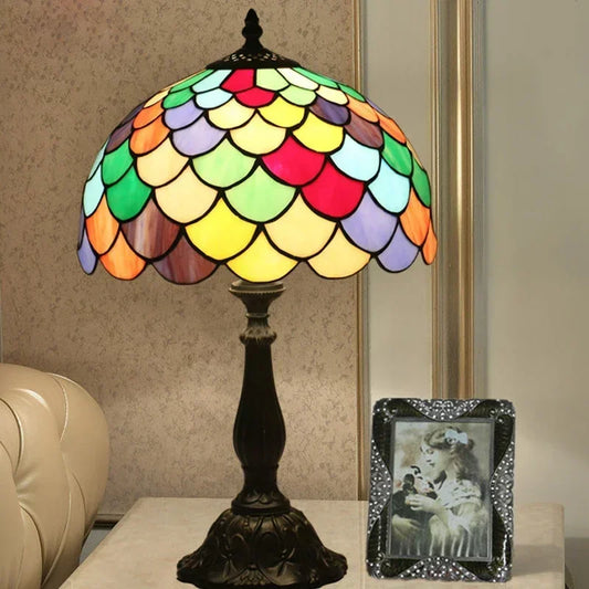A Tiffany-style table lamp with a multicolored stained glass dome shade and a dark iron base, shown on a side table next to a framed photo.