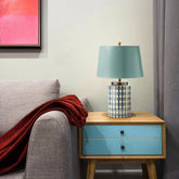 A blue ceramic table lamp with a geometric pattern and a light blue fabric shade sits on a wooden nightstand next to a grey armchair with a red throw.