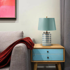 A blue ceramic table lamp with a geometric pattern and a light blue fabric shade sits on a wooden nightstand next to a grey armchair with a red throw.