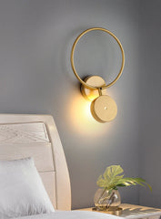 Gold wall sconce on a gray wall with a bed and plant in the foreground