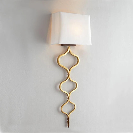 A gold wall sconce with a decorative quatrefoil chain design and a white fabric rectangular shade mounted on a light gray wall.