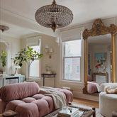 A plush pink sofa and a large, ornate gold mirror in a living room, with a crystal orb chandelier illuminated above.