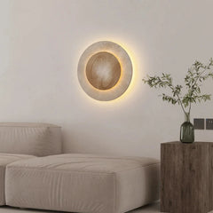 A contemporary round stone wall light with a warm LED glow, positioned on a white wall above a beige upholstered ottoman, with a wooden side table holding a vase of greenery.