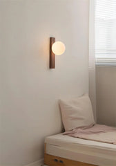 A cozy bedroom scene featuring a wood and frosted glass sphere wall sconce mounted above a bed with a white pillow and pink throw.