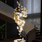 A cascading chandelier with gold floral and leaf accents and clear crystal droplets, illuminated and hanging near a modern staircase.