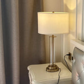 A clear glass cylinder table lamp with a brushed gold base and accents sits on a white nightstand next to a grey curtain.