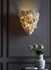 Decorative wall light fixture on a dark paneled wall with a vase and flowers on a table below.