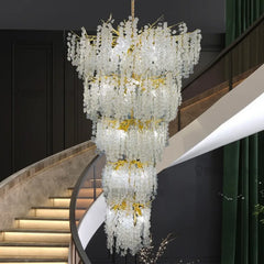 A grand, multi-tiered gold chandelier with cascading clear crystals hangs in a modern interior with a curved staircase.