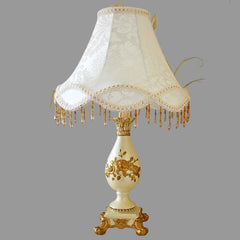 Decorative table lamp with white lace shade and gold base against a floral wallpapered wall.