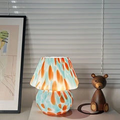 Colorful lamp with a bear figurine on a surface, with a framed picture and blinds in the background.
