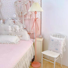 Pink floor lamp with a pleated fabric shade and a large pink bow accent, standing next to a bed with ruffled bedding.