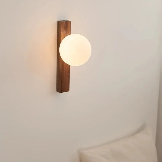 A minimalist wall sconce with a rectangular dark wood base and a glowing white spherical frosted glass shade, mounted on a white wall.