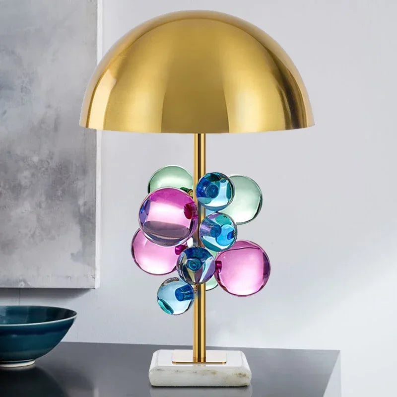 A modern table lamp with a brass finish stem, colorful glass orbs, and a white marble base, set against a neutral background.