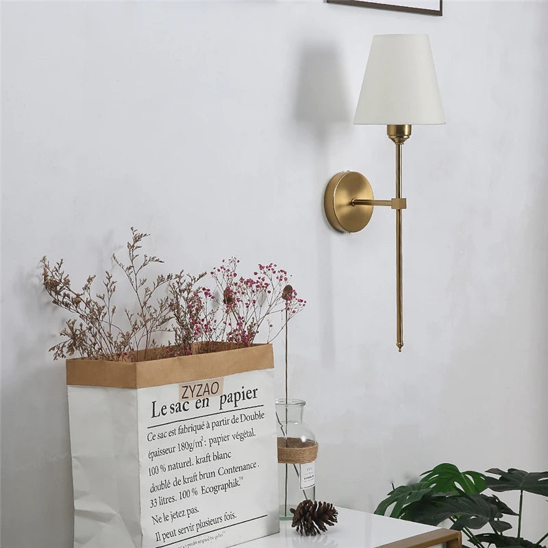 A brushed gold wall sconce with a white fabric shade mounted on a white wall above a table with dried flowers.