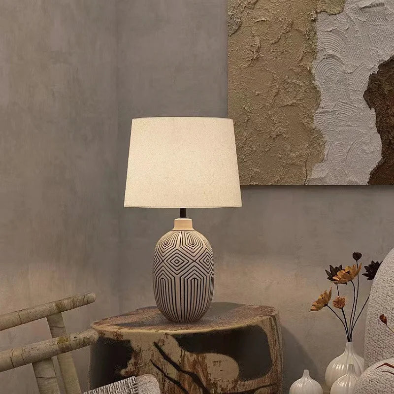 Close-up of a ceramic table lamp with a distinctive geometric pattern on its base, topped with a cream fabric shade, on a textured wooden surface.
