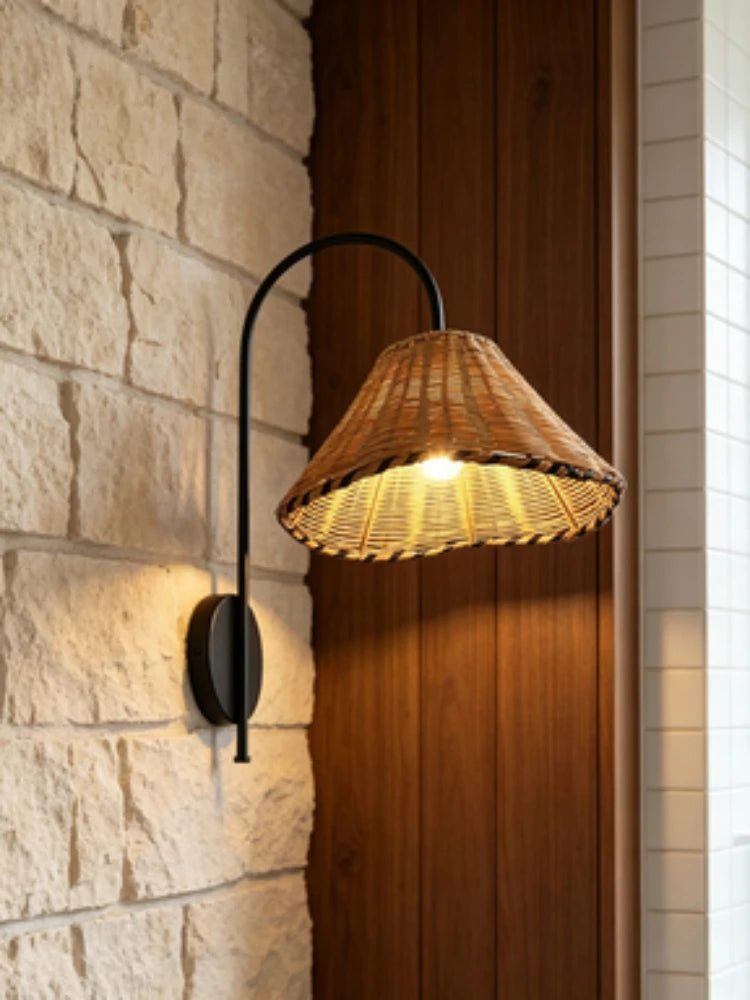 Black metal arm wall sconce with a woven rattan cone shade mounted on a stone wall next to a wooden panel.