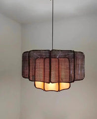 A dark-toned rattan woven tiered pendant light hanging in a dimly lit room, emitting a warm orange glow from within.