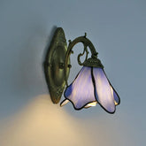 A Tiffany-style wall sconce with a purple stained glass shade shaped like a flower, mounted on a distressed gold metal arm against a textured wall.