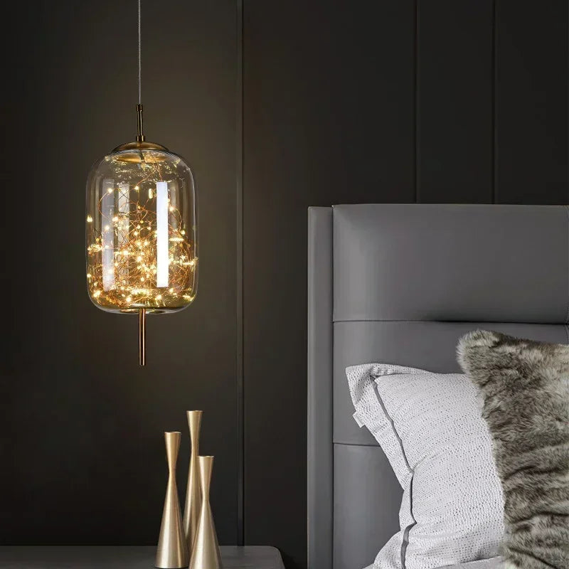 A glass cylinder pendant light filled with warm LED lights, hanging next to a modern bed.