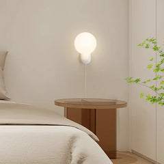 A minimalist wall sconce with a white globe shade mounted on a wall next to a bed.