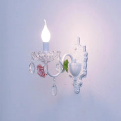 A single flame-tip bulb illuminates a white wall sconce decorated with a pink ceramic rose and clear crystal droplets.