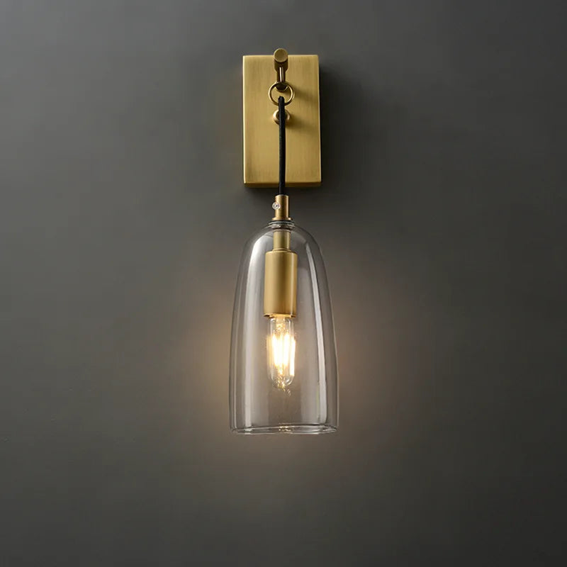 Front view of a gold and clear glass bell wall sconce against a dark gray wall, light illuminated.