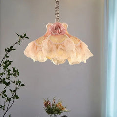 A fabric pendant light with a pink rose accent and ruffled shades hangs in a softly lit room with greenery in the foreground.