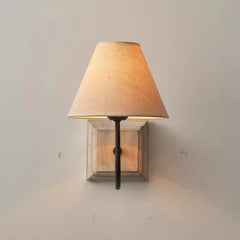A wall sconce with its light on, illuminating a light beige fabric cone shade and casting light upwards and downwards. The fixture has a distressed white wood base and a dark metal arm.