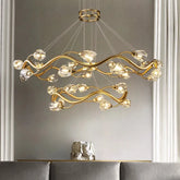 Gold wavy crystal chandelier with multiple tiers and cascading crystal accents, illuminated against a neutral wall.
