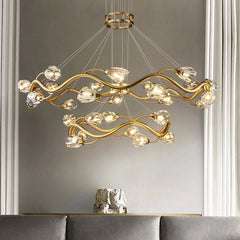 Gold wavy crystal chandelier with multiple tiers and cascading crystal accents, illuminated against a neutral wall.