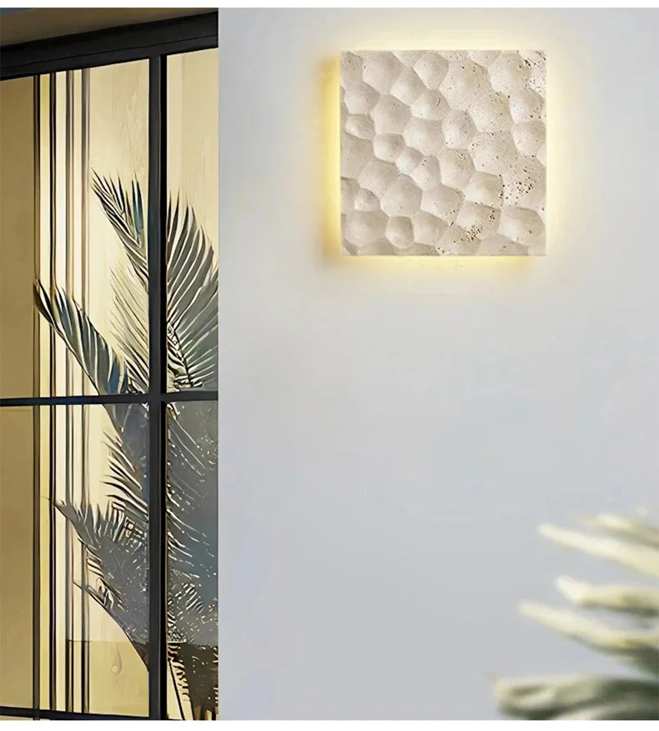 A light stone textured wall sconce mounted on a white wall next to a large window with palm leaves visible inside.