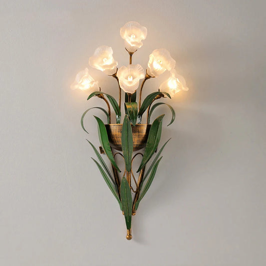 Gold floral wall sconce with clear glass flower shades and green leaf accents against a white wall.